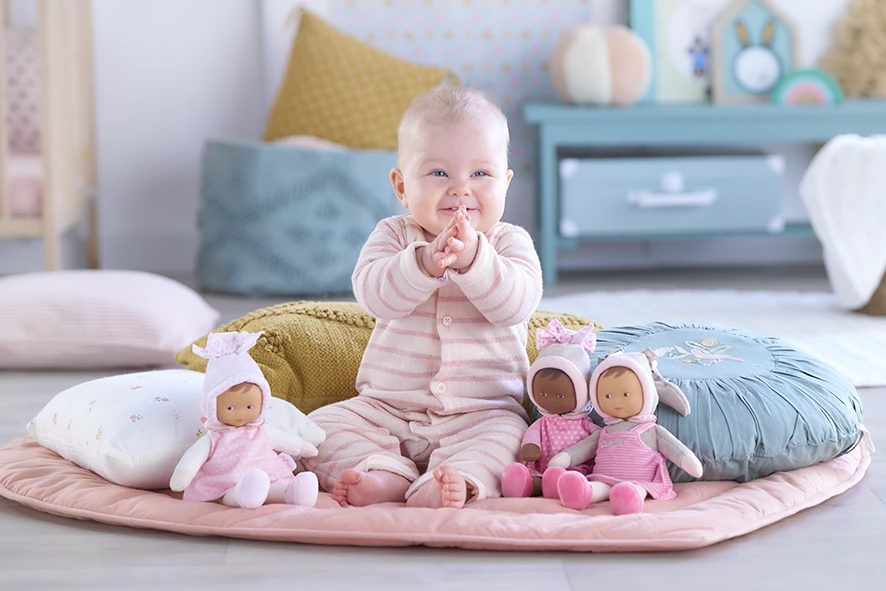 Bébé qui rigole et se frotte les mains avec à ses pieds des doudous